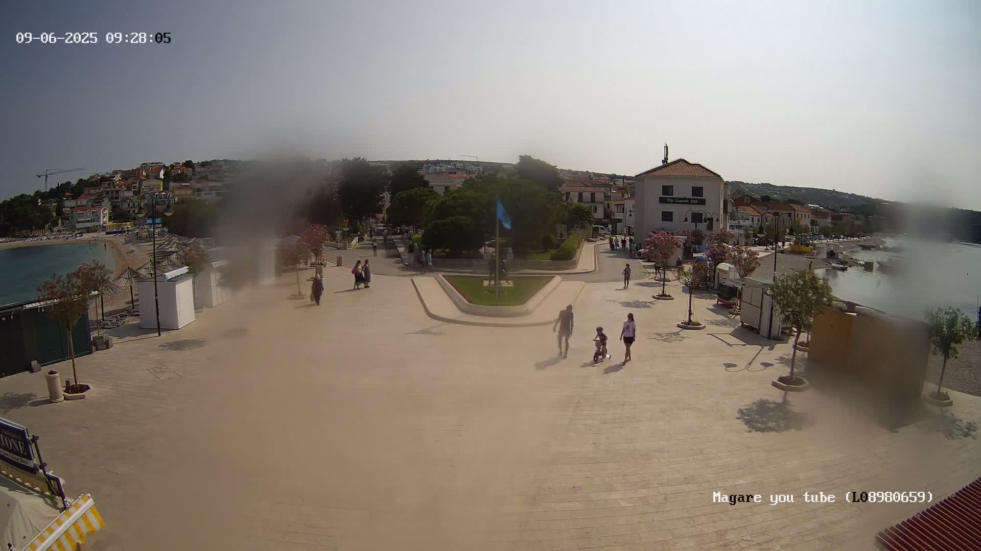 A hazy, sunny day reveals a seaside town square with a few people walking, a small beach, and buildings lining the waterfront.