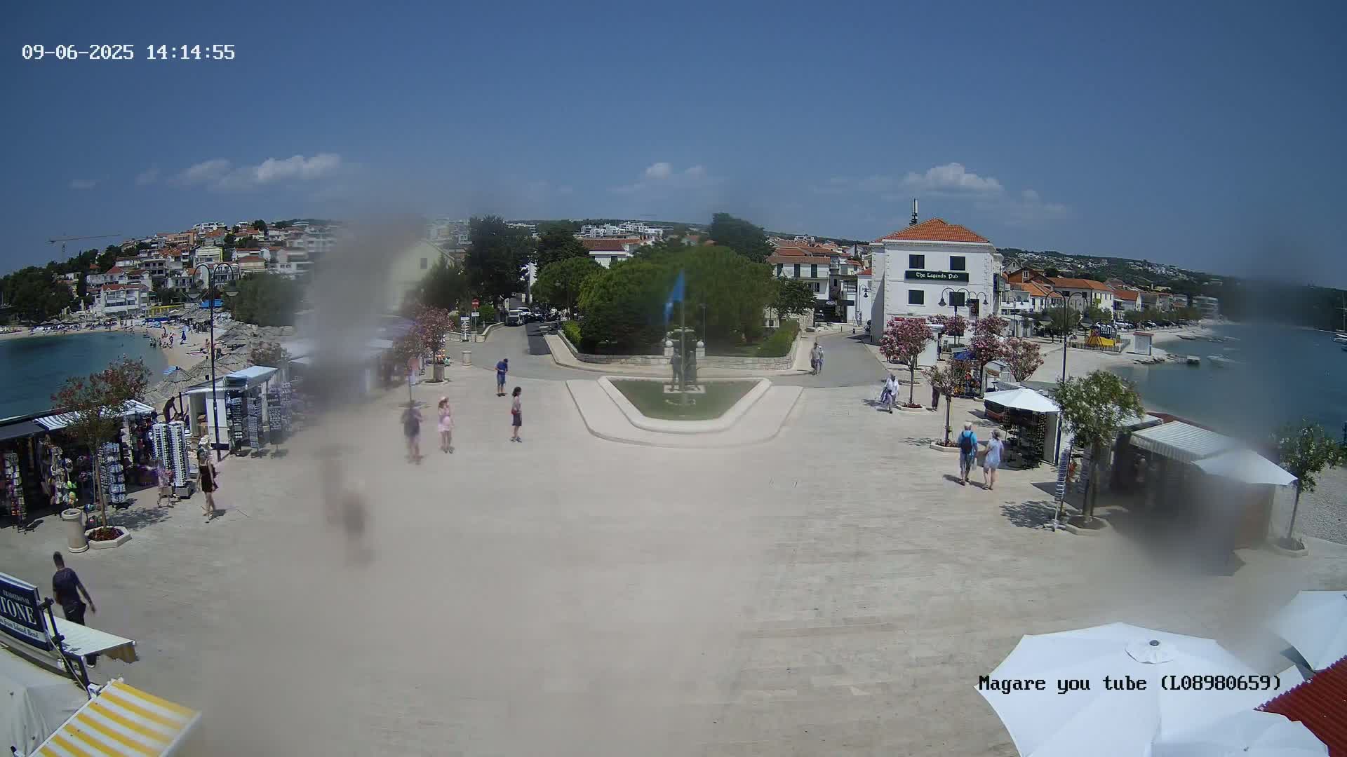 A town square by the sea on a sunny day, featuring a central fountain, several small shops, and people walking around.