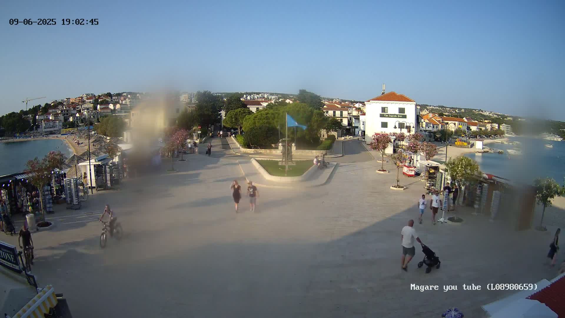 A seaside town square with people walking, vendors selling goods, and a calm body of water in the background under a clear, sunny sky.