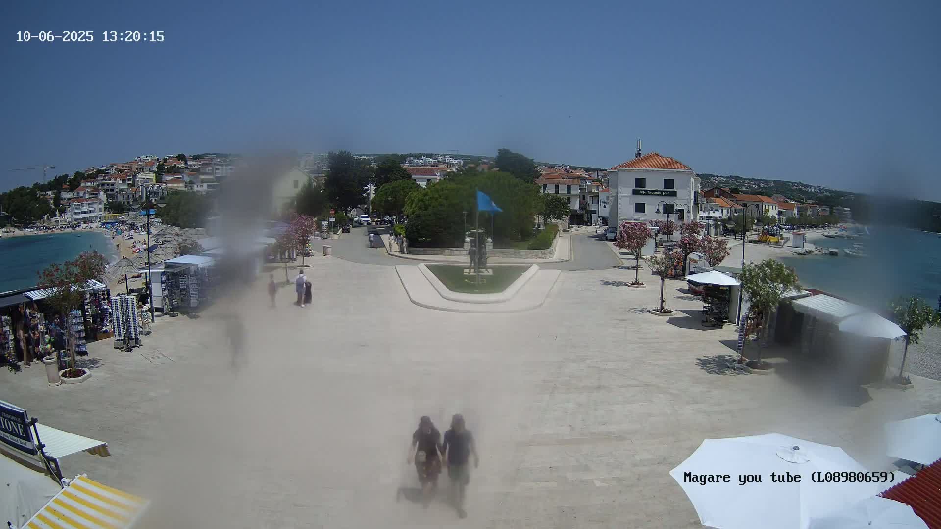 A seaside town square on a sunny day, with a monument in the center, small shops and umbrellas lining the perimeter, and people walking around.
