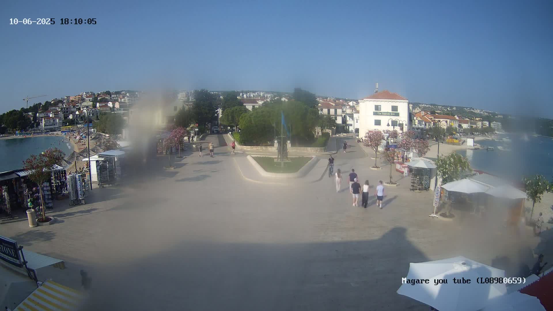 A hazy, sunny day reveals a seaside town square with people walking, souvenir stands, and a beach in the background.