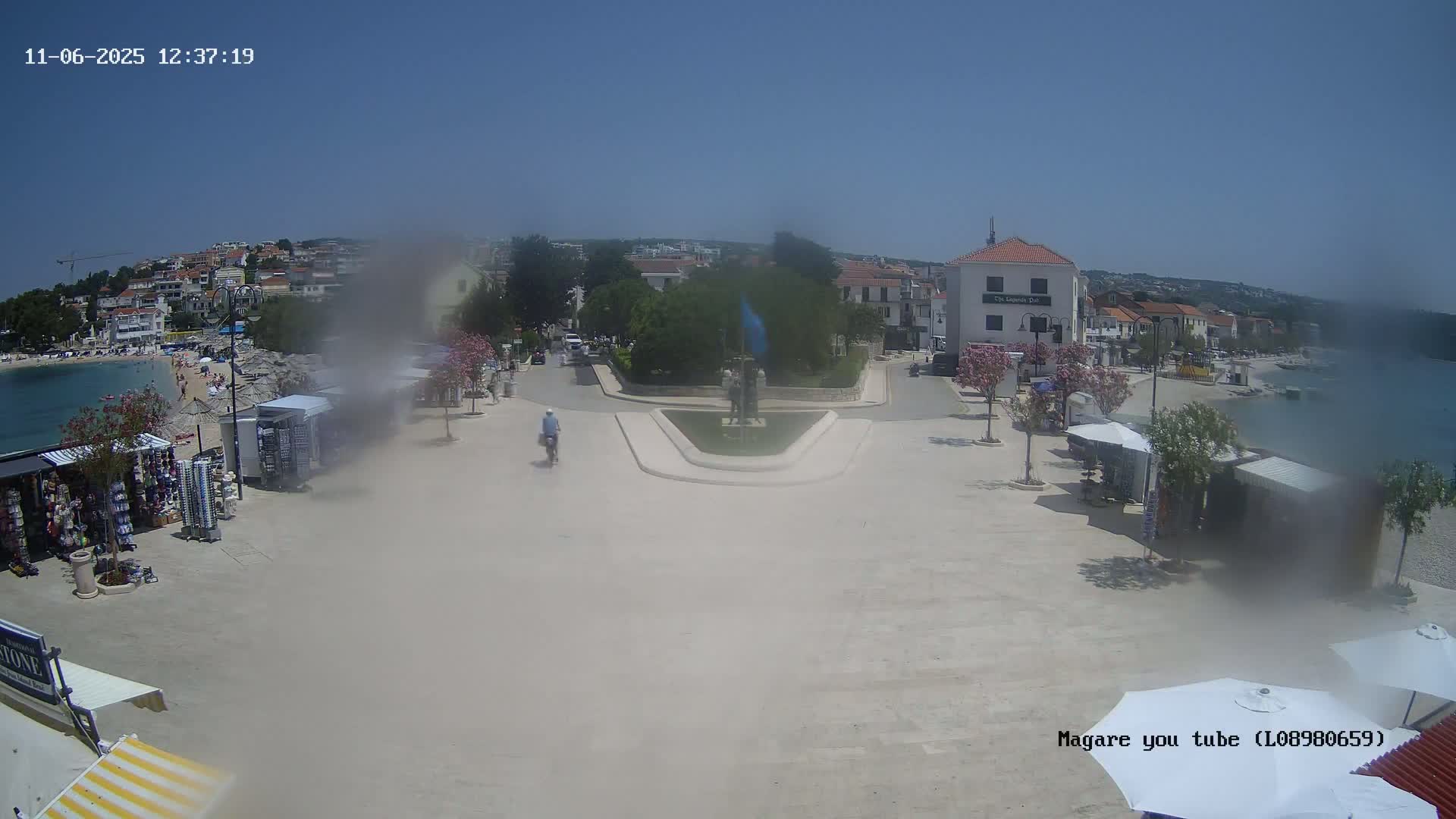 A hazy, sunny day reveals a town square by the sea, with a monument at its center, shops lining its perimeter, and a cyclist riding through the plaza.