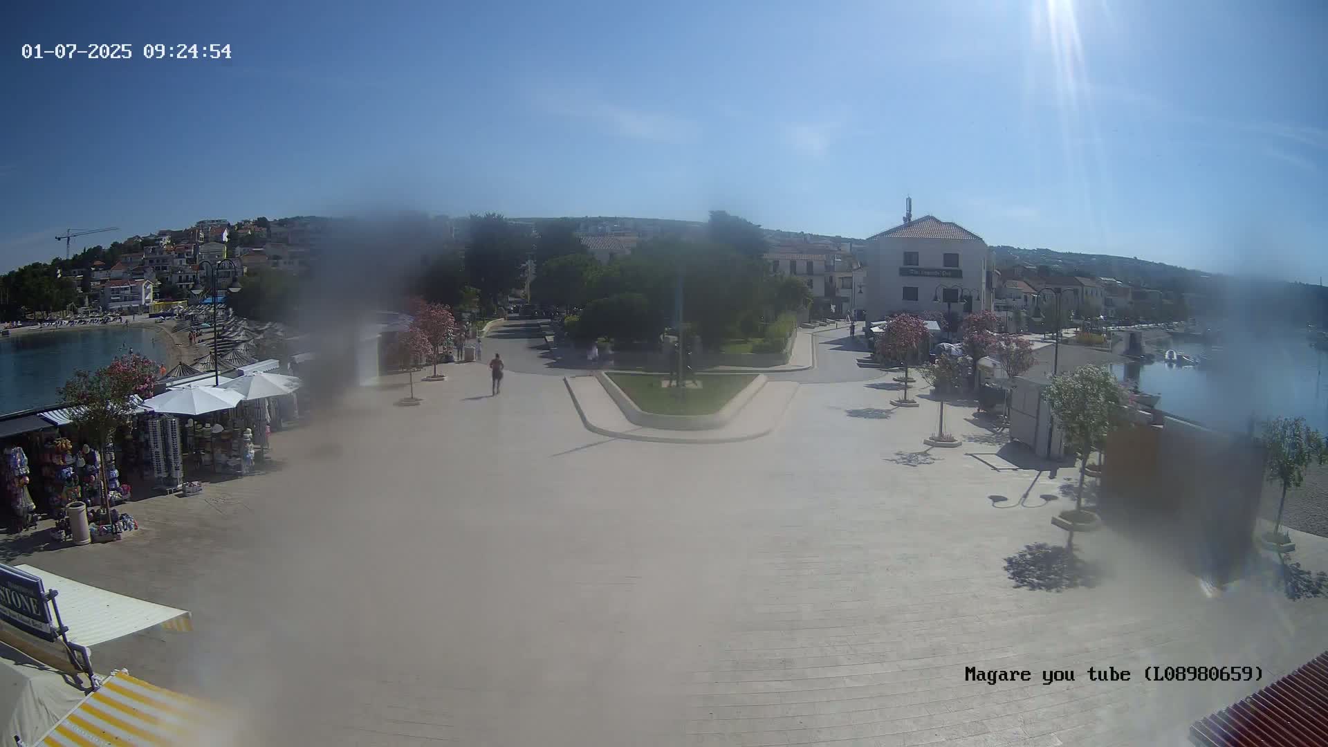 A hazy sunny day overlooking a town square near a calm body of water, with several people walking and shops with umbrellas set up along the waterfront.