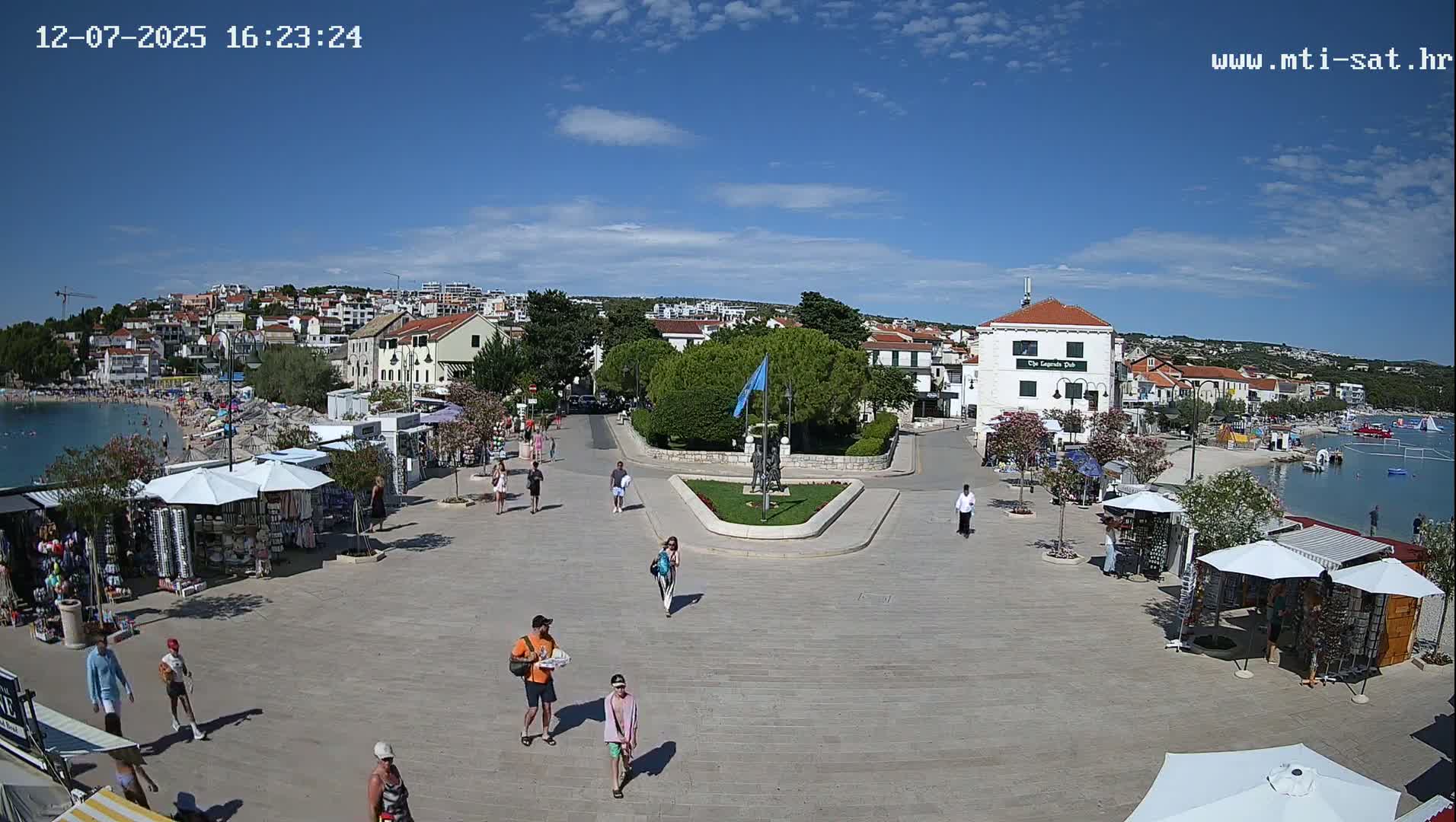 A hazy sunny day overlooking a town square near a calm body of water, with several people walking and shops with umbrellas set up along the waterfront.