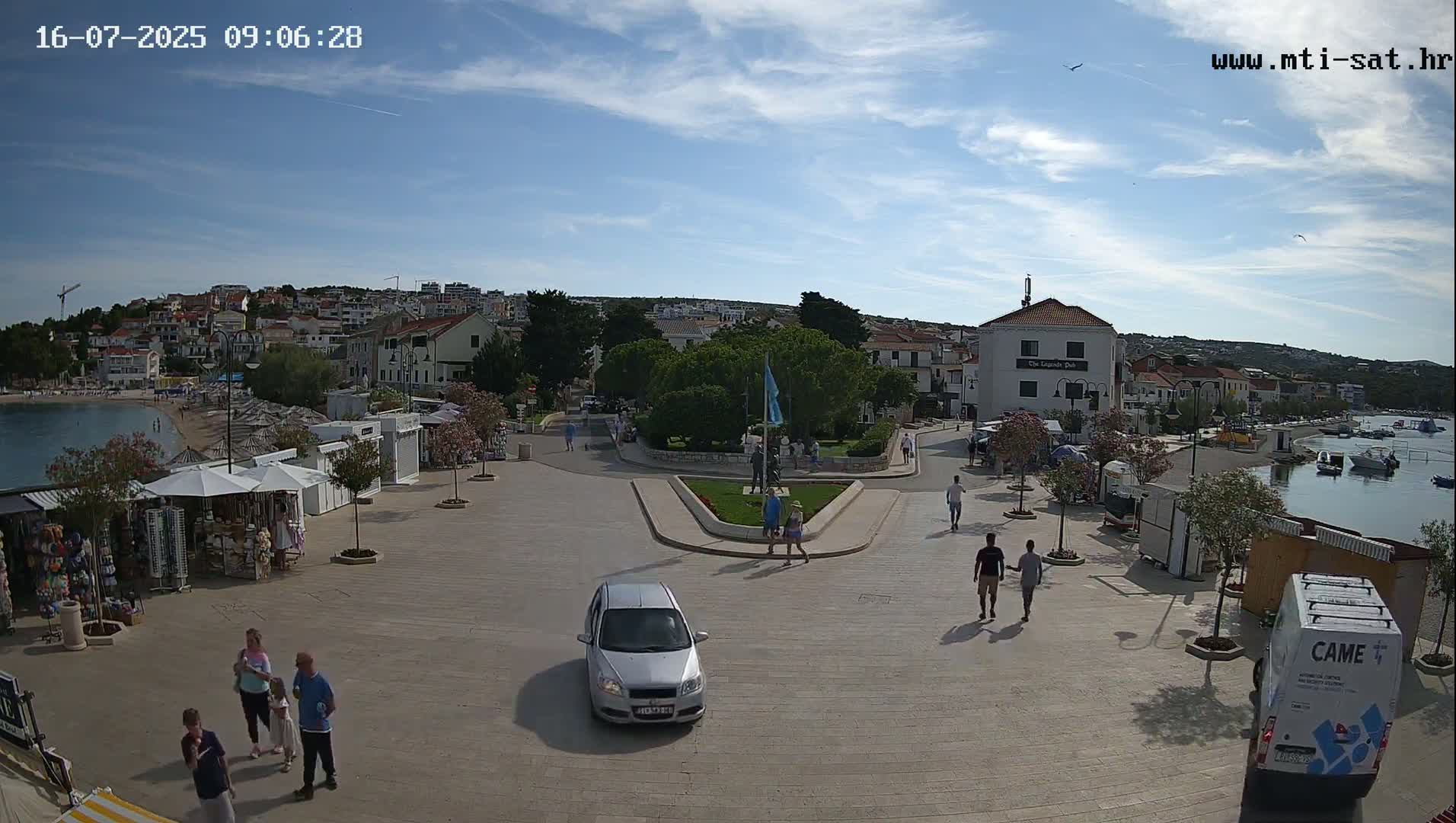 A seaside town square with a few pedestrians and cars, under a mostly sunny sky.