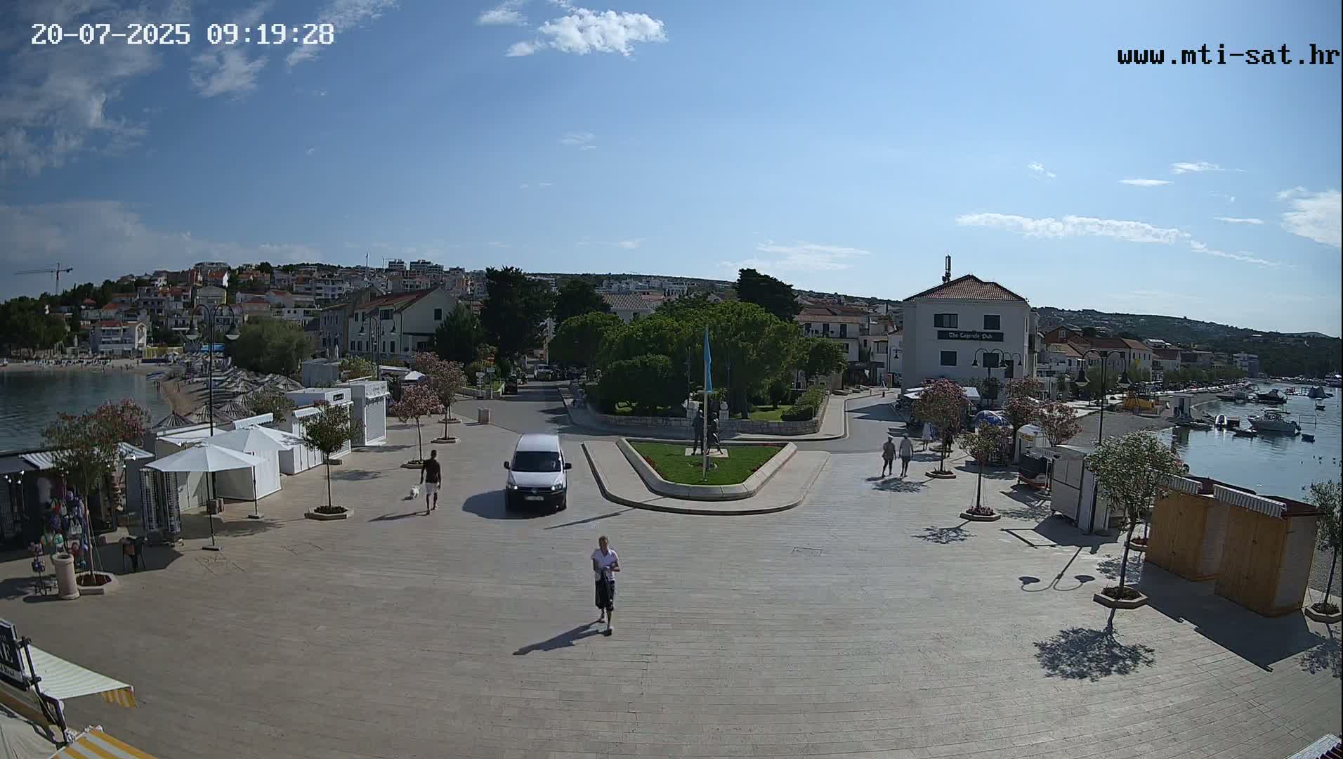 A sunny day reveals a town square overlooking a calm harbor, with several people walking, a white van parked, and small shops lining the edges.