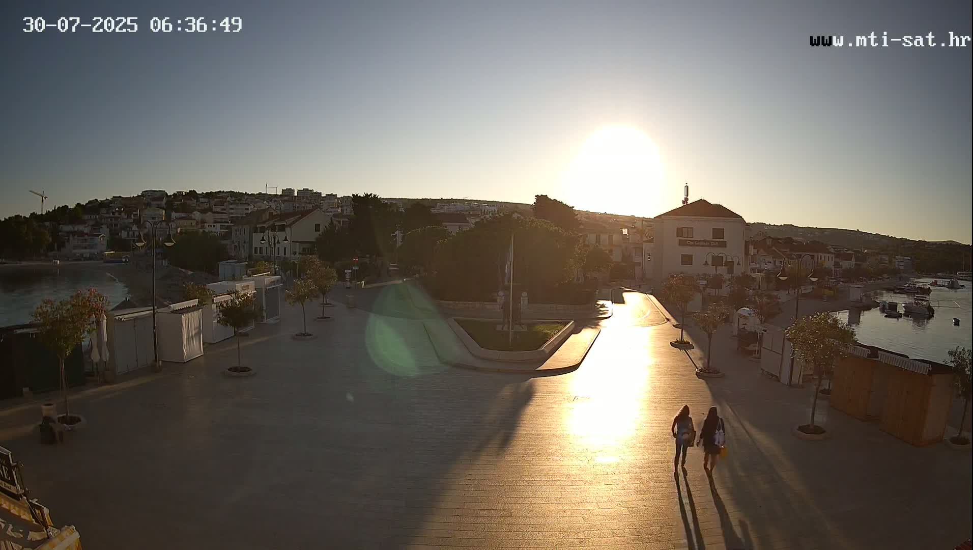 A sunny day reveals a town square overlooking a calm harbor, with several people walking, a white van parked, and small shops lining the edges.