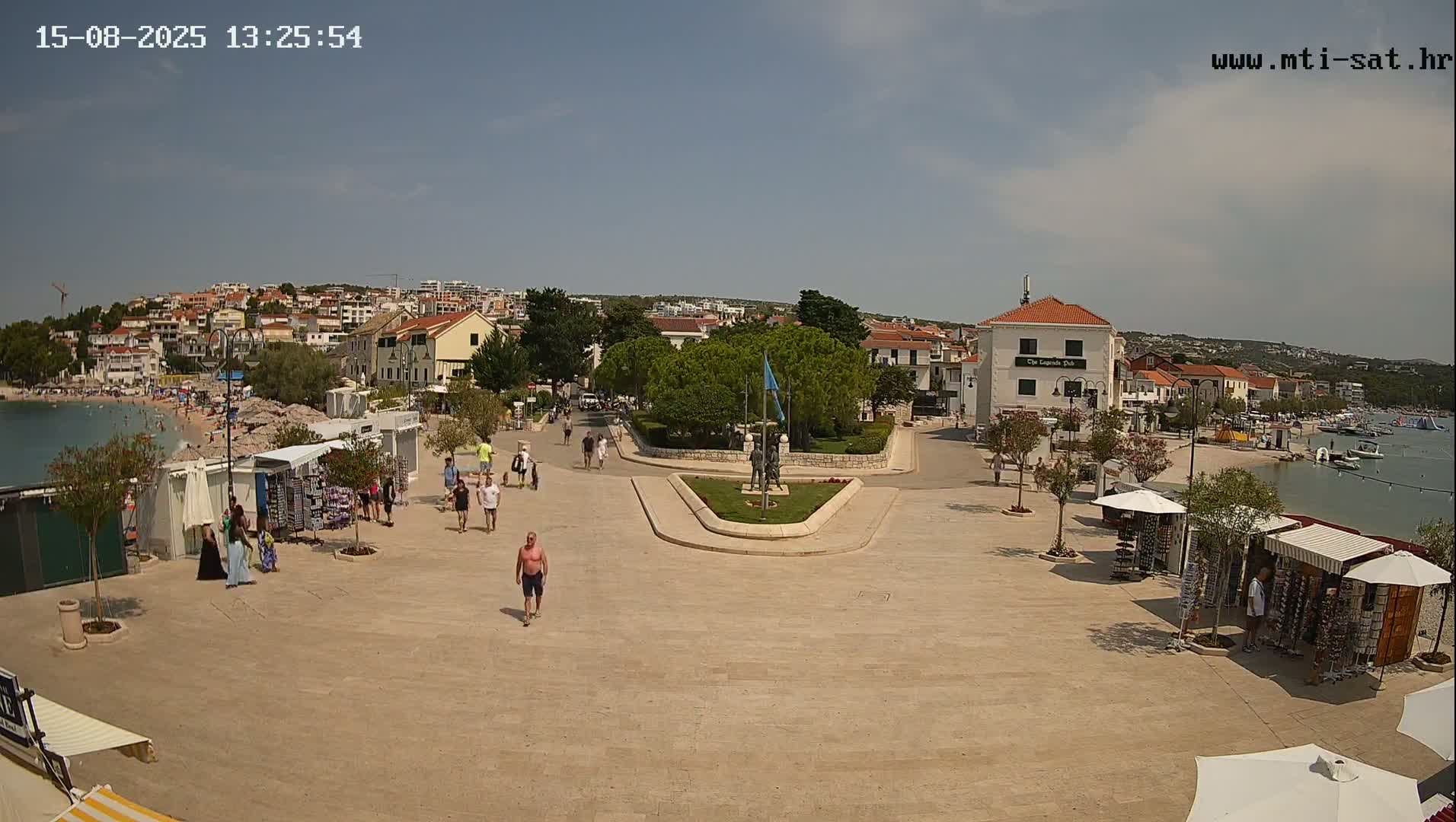 A sunny seaside town square with a few people walking around, some small shops, and a calm body of water with boats visible in the background.