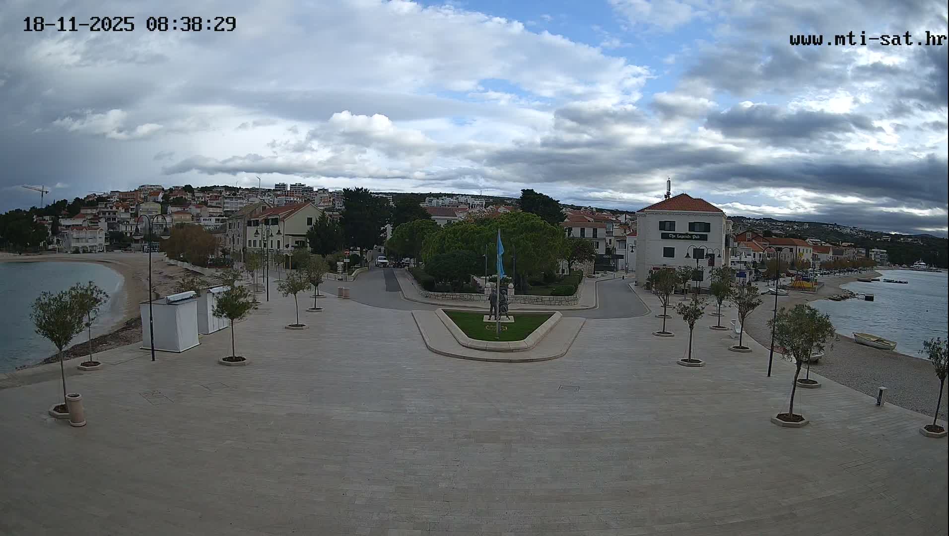 A sunny seaside town square with a few people walking around, some small shops, and a calm body of water with boats visible in the background.