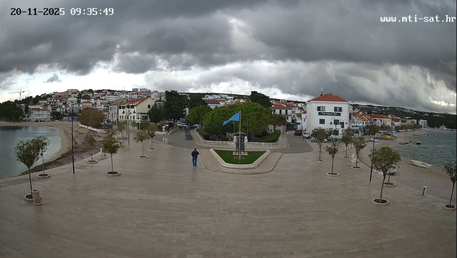 A wide-angle view captures a tranquil coastal town with a sandy beach, a bay dotted with boats, and hillside buildings, all under a heavily overcast and stormy sky, as a lone person walks across a paved promenade towards a roundabout with a statue and a blue flag.