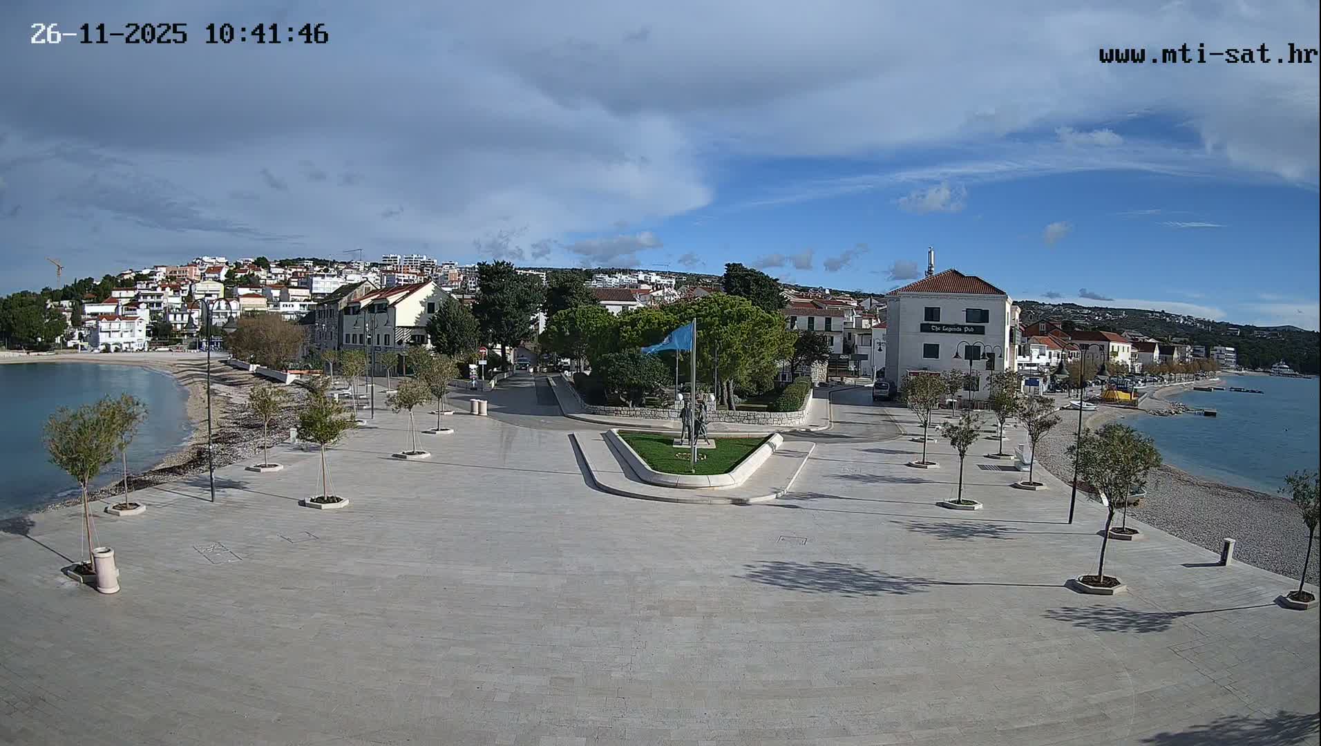 A seaside town is depicted from an elevated perspective, showcasing a calm bay with a pebble beach and a wide promenade lined with young trees, extending to a central square with a statue, all set against a backdrop of white-roofed buildings climbing the surrounding hills under a partly cloudy sky with periods of sunshine.