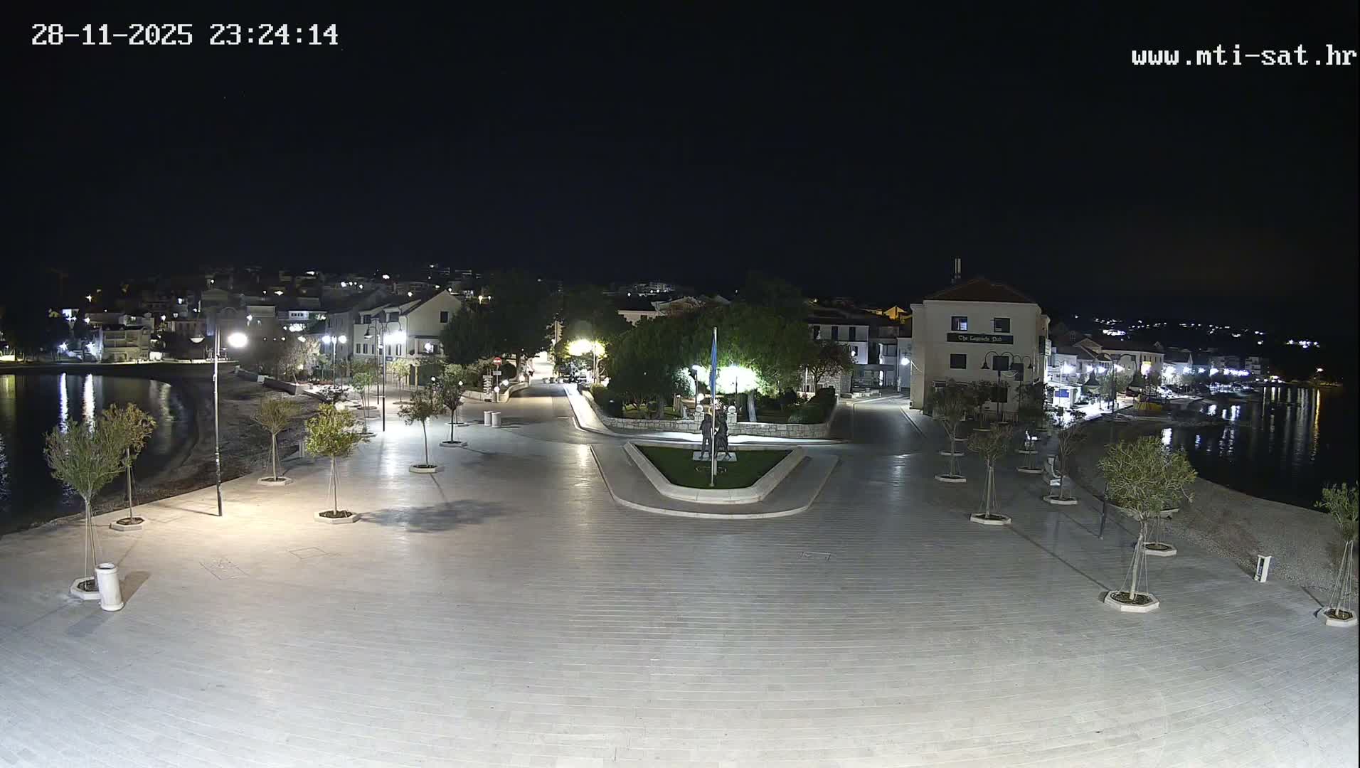 A clear night view shows a brightly lit paved waterfront promenade, lined with small trees and buildings, bordering a calm body of water reflecting the city lights under a dark, starry sky.