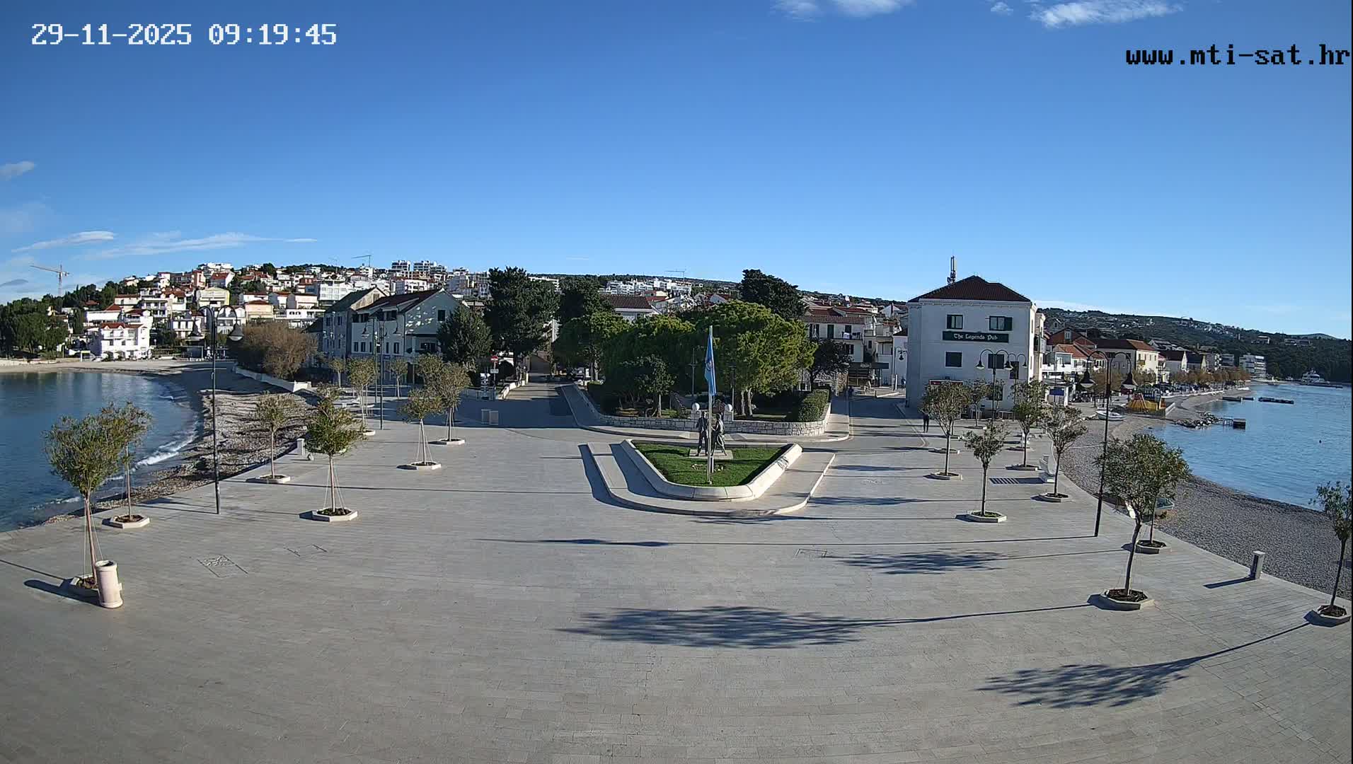 The image shows a sunny coastal town with a wide paved promenade, a pebbly beach, and calm blue sea on the right, a central green area with a flag and statue, and various buildings nestled into a hillside in the background, all under a clear blue sky.