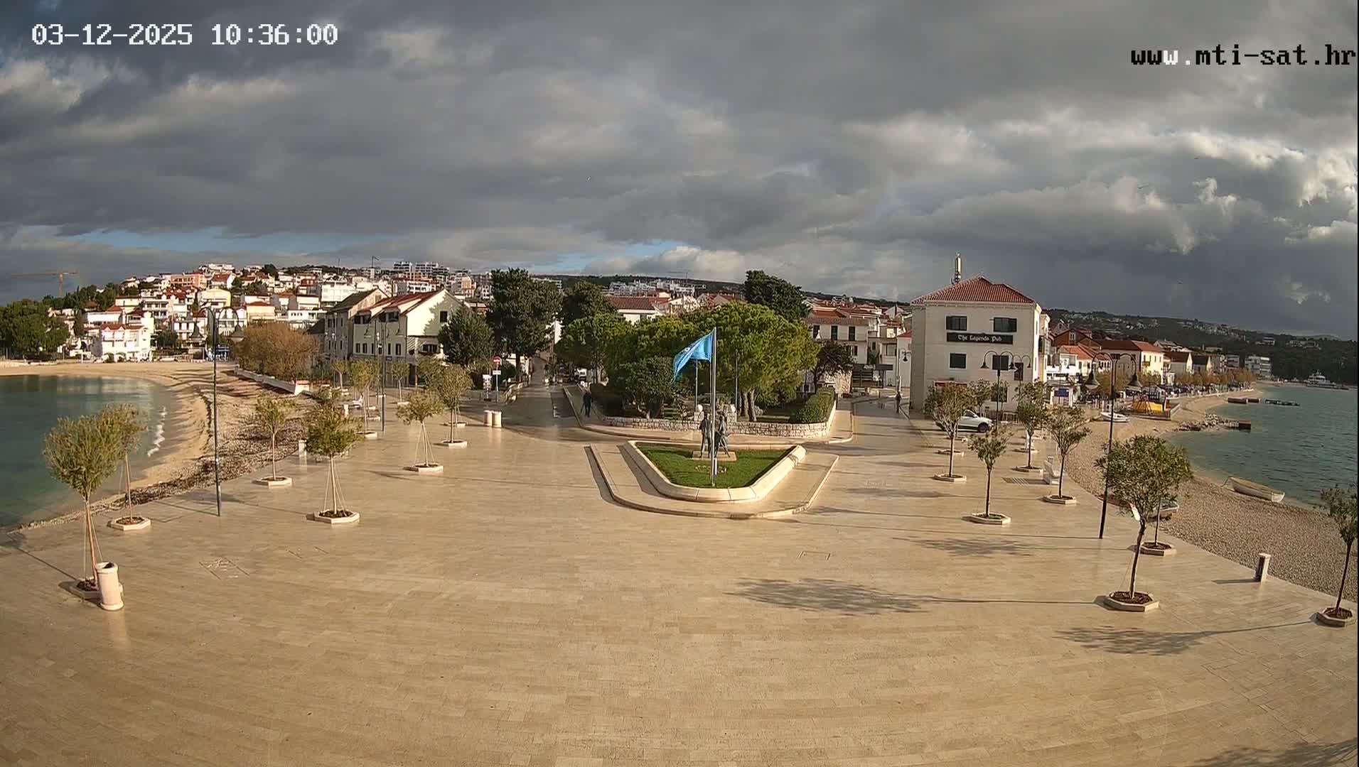 A wide paved promenade with small trees lines a pebble beach and calm sea in a coastal town, featuring a central green island with a statue and blue flag, all under a heavily overcast sky with some lighter patches.