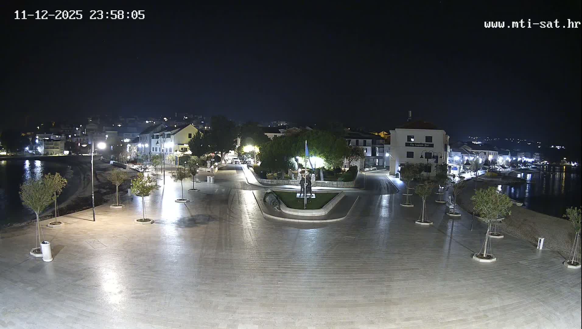 A nighttime view reveals a well-lit coastal town promenade with wet, reflective pavement, buildings lining the waterfront, small trees, and a central roundabout featuring statues and a flagpole, all under a clear sky.