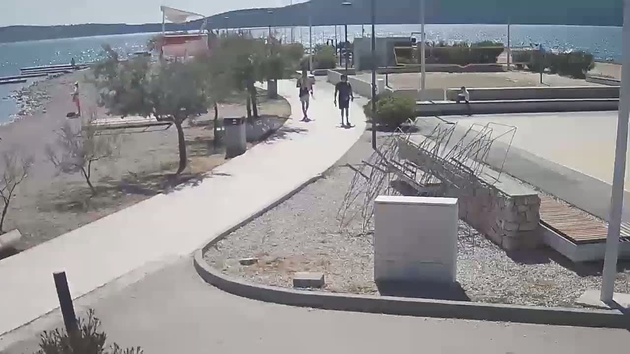 A paved walkway winds through a seaside area with sparse vegetation, several people walking, and a calm body of water visible in the background under a sunny sky.