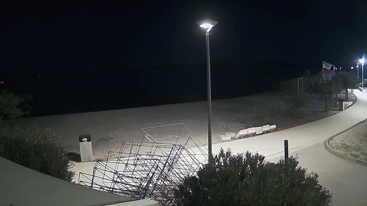 A nighttime view of a beach with a paved walkway, several lights, and a pile of metal frames near the sand.