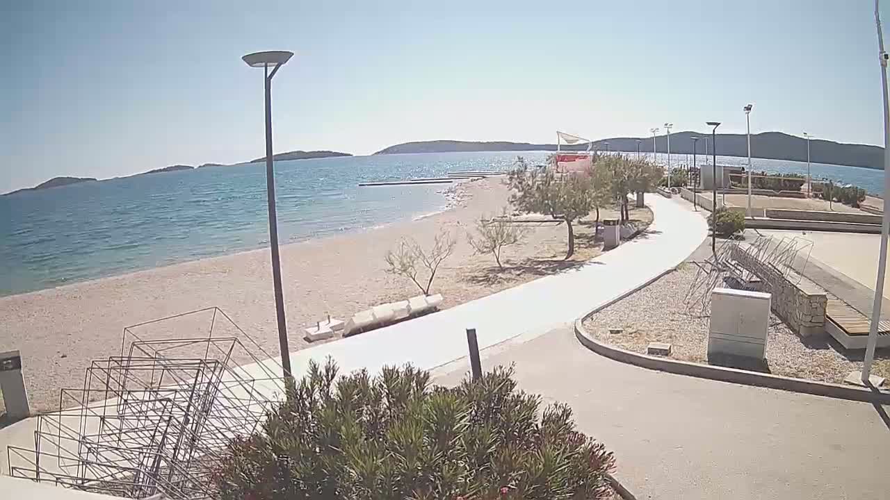 A sandy beach with a paved walkway leads to a calm, blue sea under a clear, sunny sky, backed by low hills.