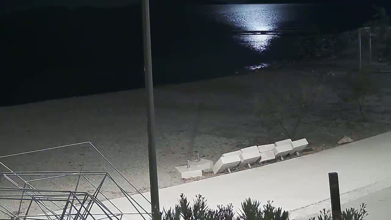 A moonlit beach at night shows a paved walkway near the water's edge with several concrete blocks along it, and a metal structure lying on the sand.