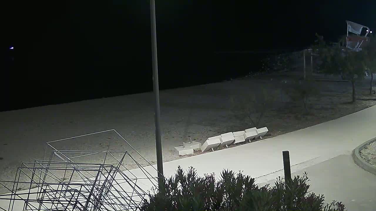 A nighttime beach scene shows a light-colored paved path, a metal sculpture, several white blocks, and low shrubs near a dark body of water under a dark sky.