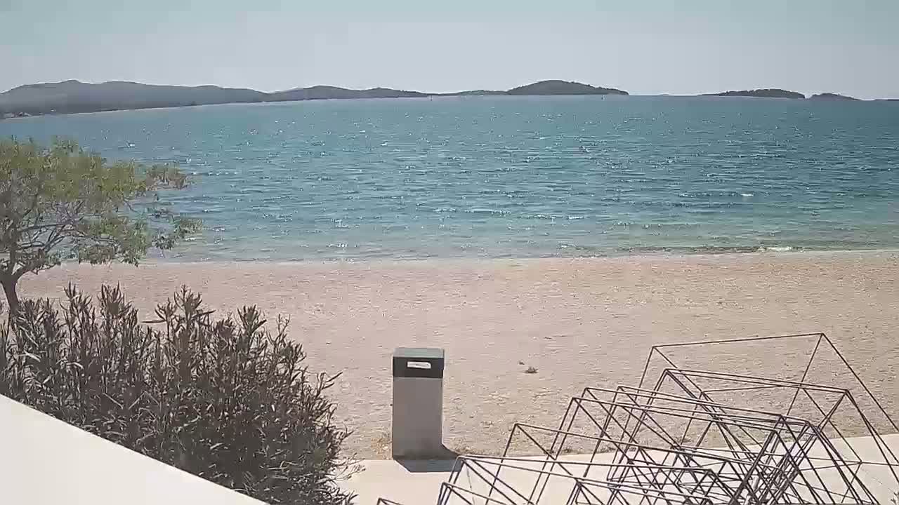 A sandy beach with a calm, blue sea extends to distant hills under a sunny sky, featuring a trash receptacle and a metal sculpture in the foreground.