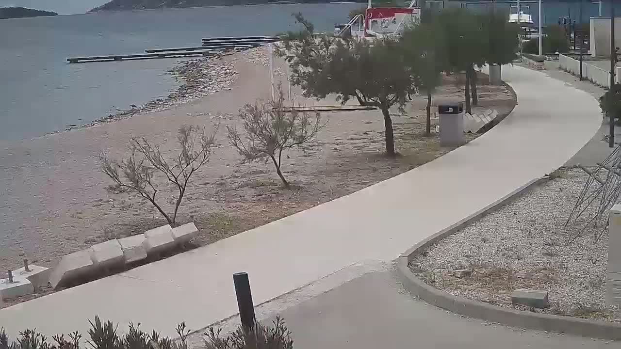 A paved walkway curves past a sandy beach and calm water toward a distant shoreline under an overcast sky.