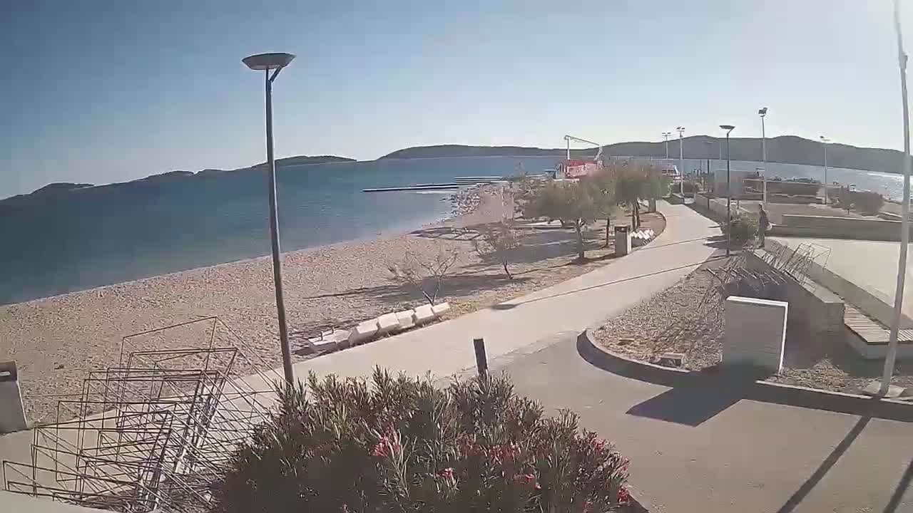 A sandy beach with a paved walkway, surrounded by low-lying vegetation and lampposts, extends towards a calm, blue sea under a clear, sunny sky.