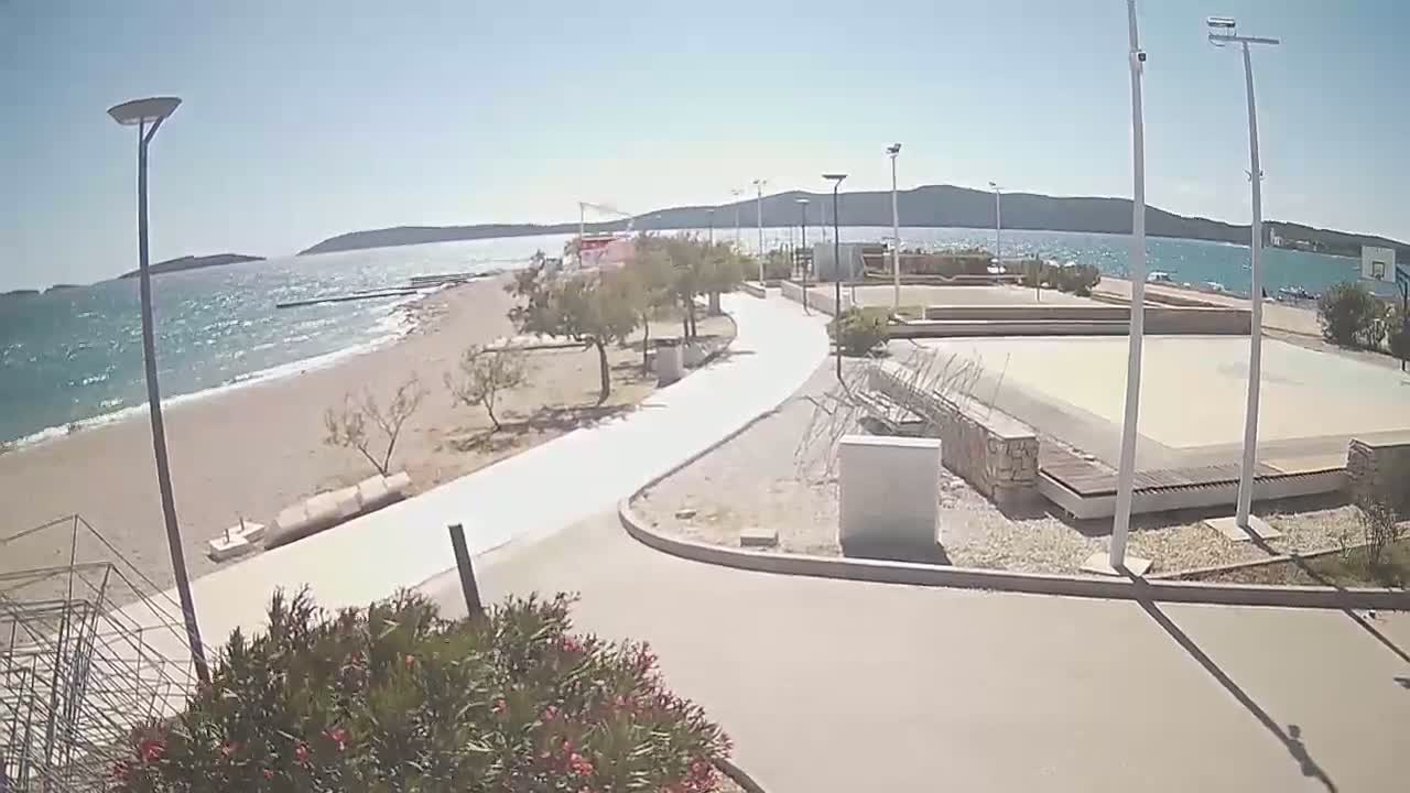 A paved walkway winds through a beachfront area with sparse vegetation, a sandy beach, calm ocean water, and an outdoor basketball court, all under a sunny sky.
