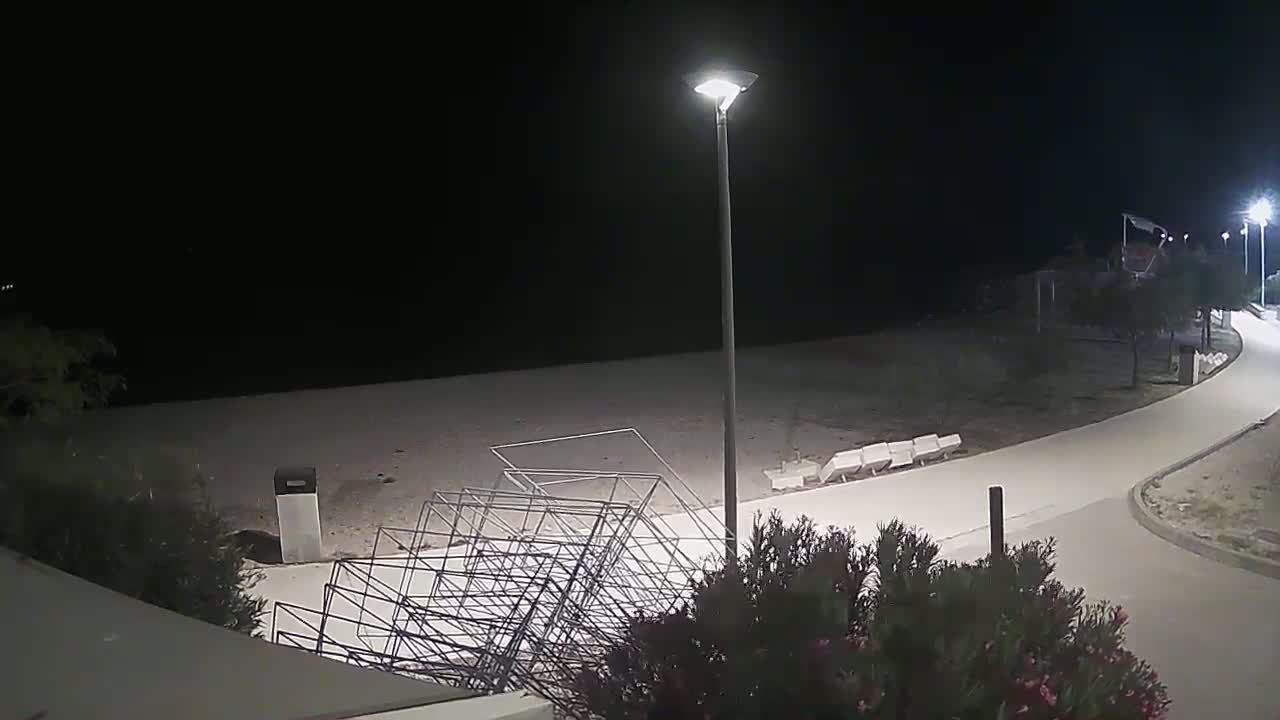 A nighttime view of an empty beach with overturned metal beach chairs, a paved walkway, and vegetation.