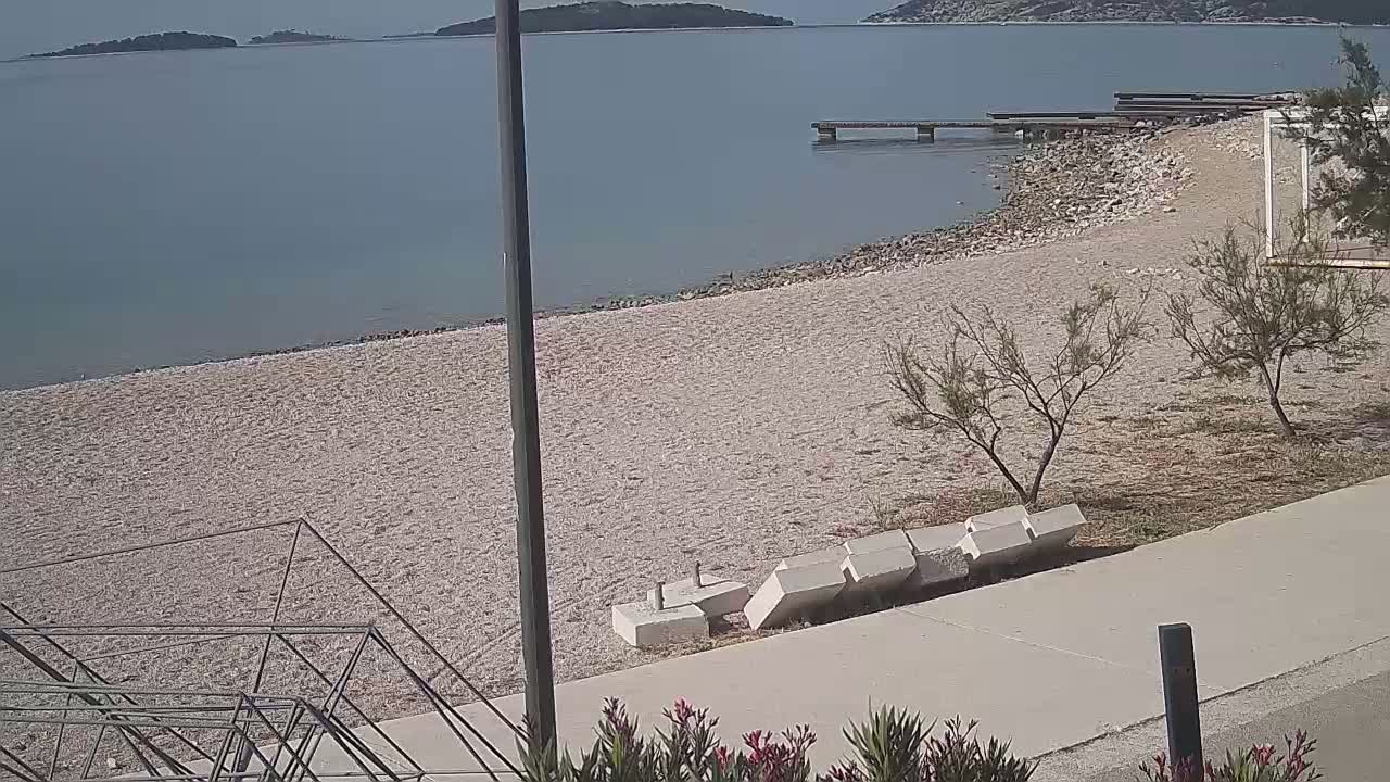 A mostly empty pebble beach stretches toward calm blue water, under a clear sky, with small shrubs and a concrete walkway bordering the sand.