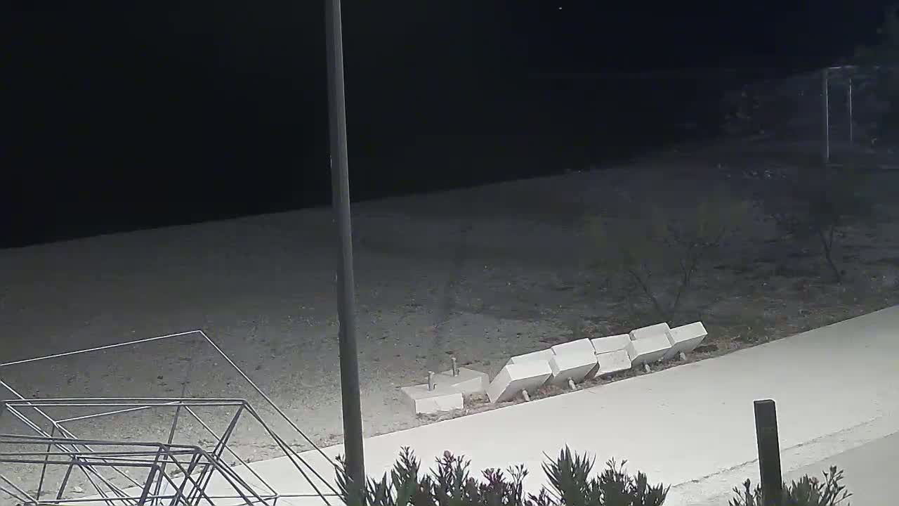 A nighttime view of a paved walkway next to a sandy area with scattered, damaged concrete blocks, and a section of what appears to be a metal shopping cart frame lying on the ground.