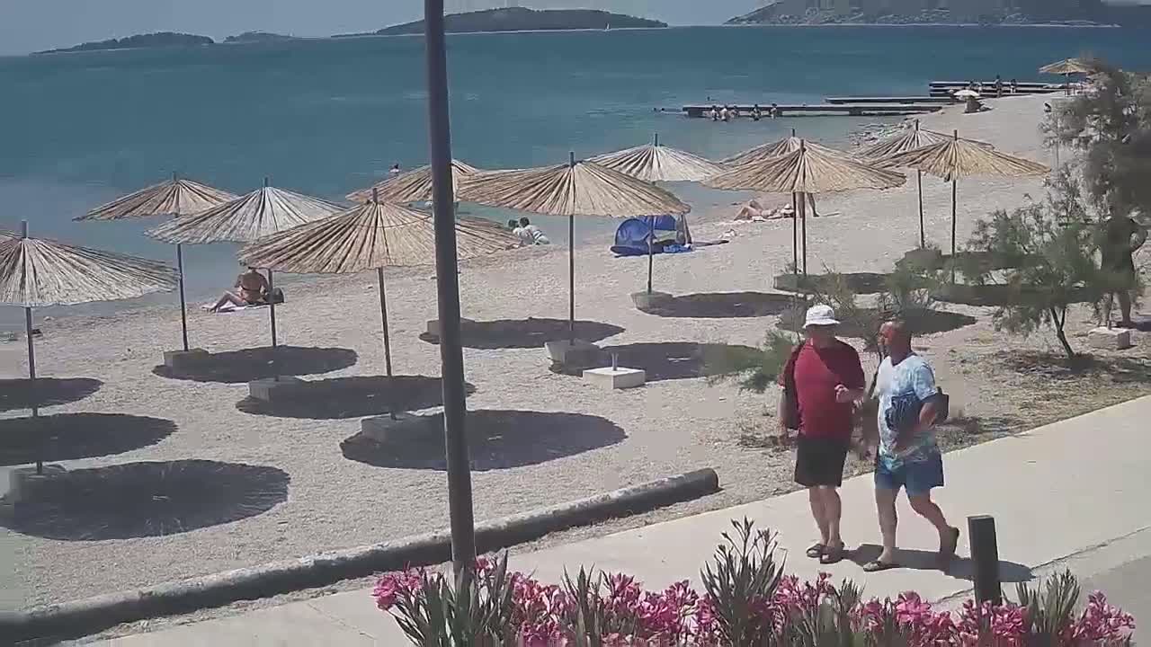 Two men walk along a paved path next to a pebble beach with straw umbrellas and scattered sunbathers under a sunny sky.