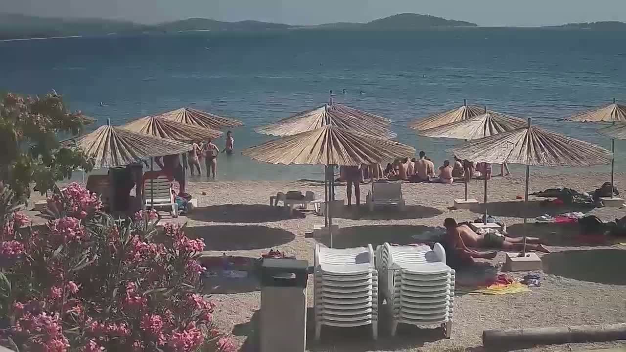 A sandy beach with numerous straw umbrellas and beach chairs is populated by people swimming and relaxing on a sunny day next to calm, blue water with distant hills visible in the background.