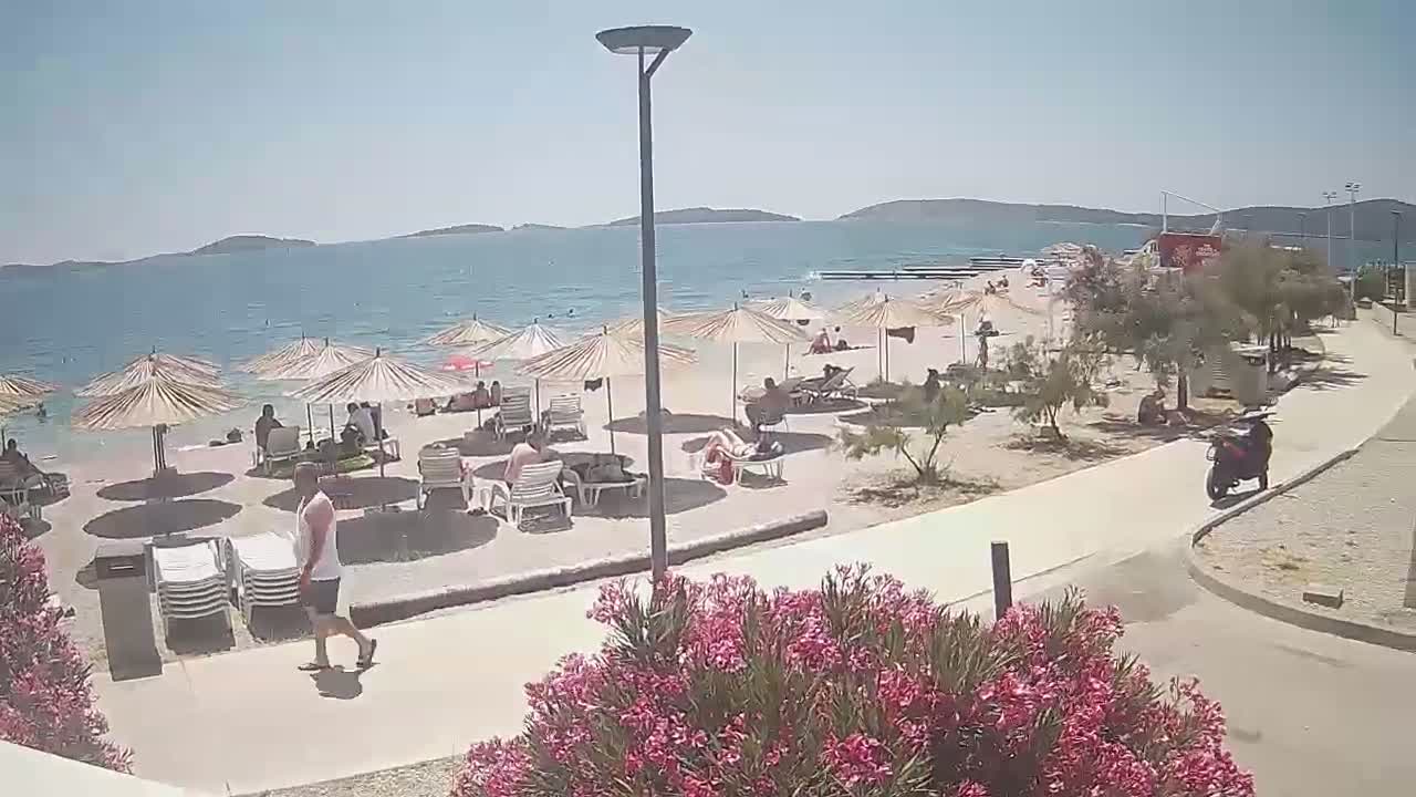 A sunny beach scene shows people relaxing under umbrellas on a sandy beach next to a paved walkway and calm blue water with islands in the distance.