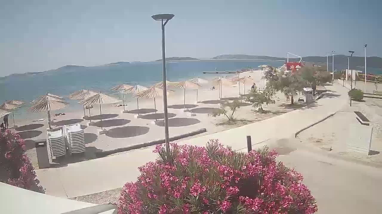 A sandy beach with numerous closed umbrellas and a few people is visible, next to a paved walkway, under a sunny sky and calm sea.