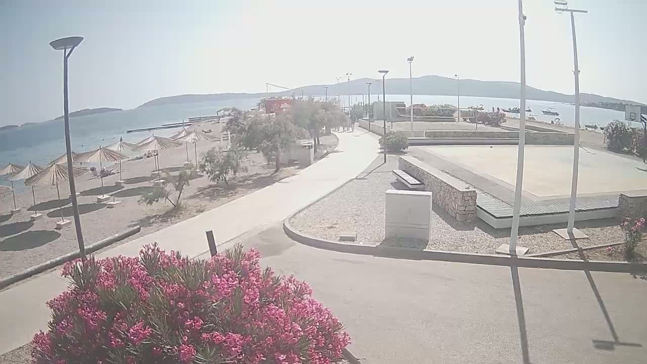 A paved walkway leads from a sandy beach with umbrellas to an outdoor basketball court, all under a bright, sunny sky.