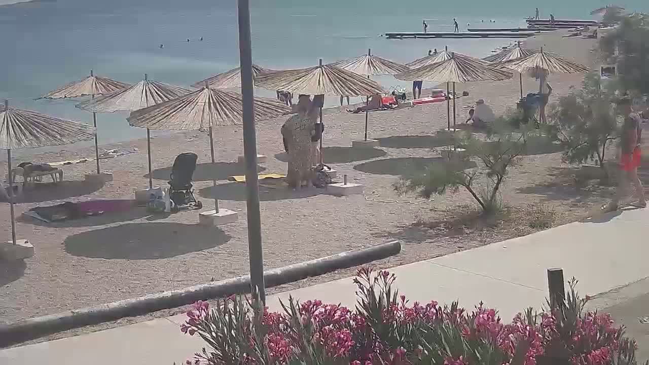 A sandy beach with straw umbrellas, several people, and a stroller is visible on a sunny day near calm blue water.