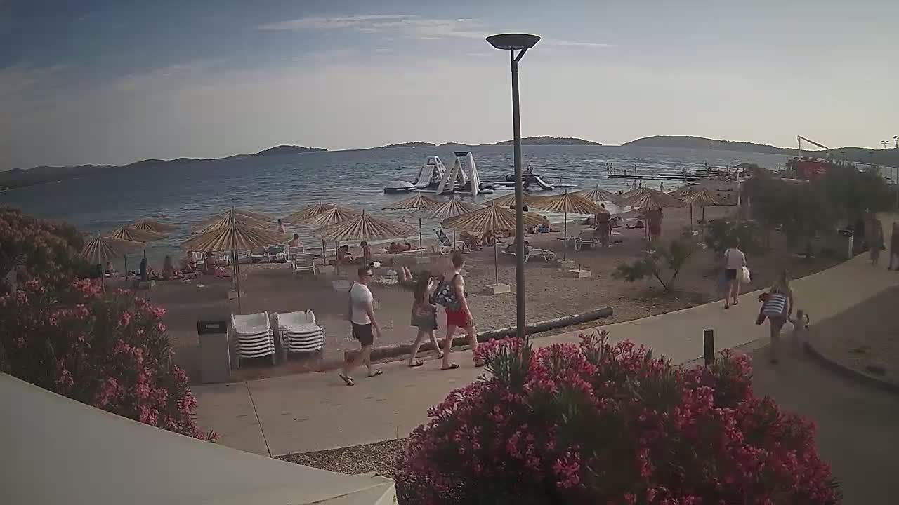 A sandy beach with numerous umbrellas and people, some in the water near inflatable water toys, on a partly cloudy day.