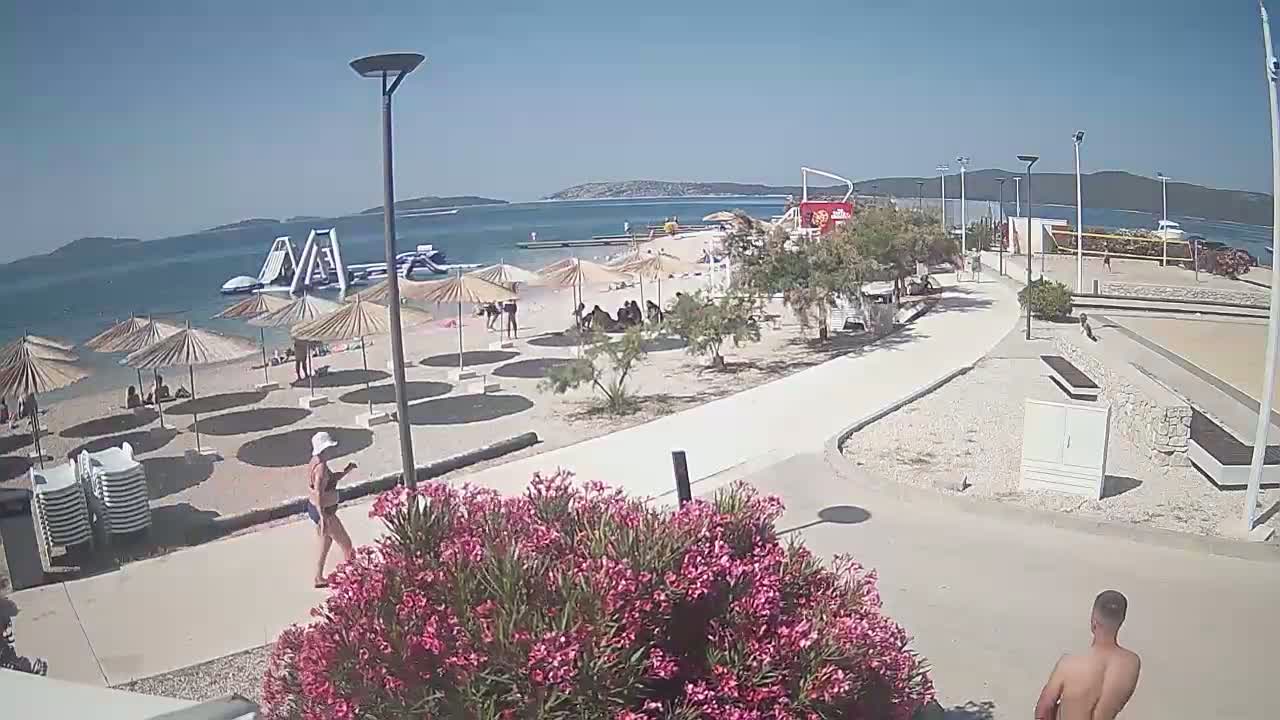 A sunny beach scene shows people relaxing under umbrellas, near inflatable water toys in the ocean, with a paved walkway leading to a volleyball court and other beachside amenities.