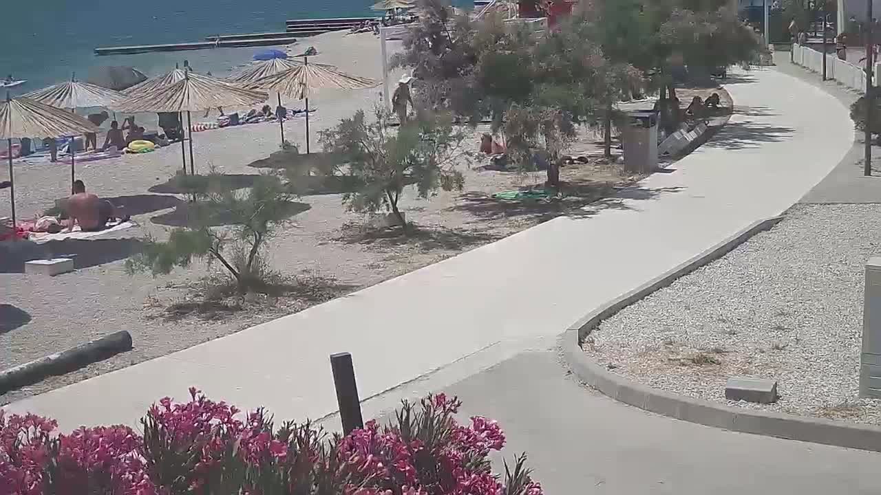 A paved walkway curves alongside a sandy beach where people are relaxing under straw umbrellas on a sunny day.