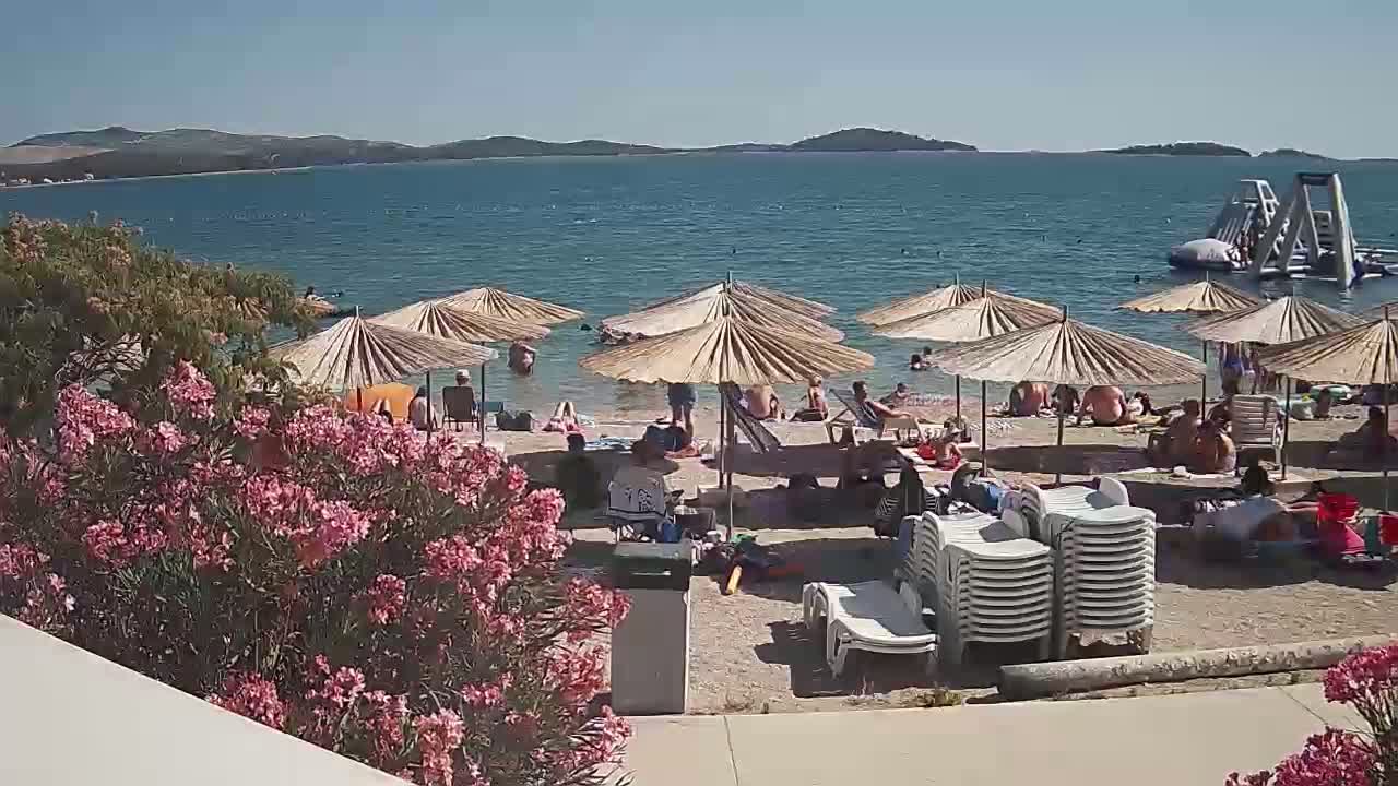 A sandy beach with many people relaxing under umbrellas near a calm body of water, on a sunny day, with an inflatable water park visible in the distance.