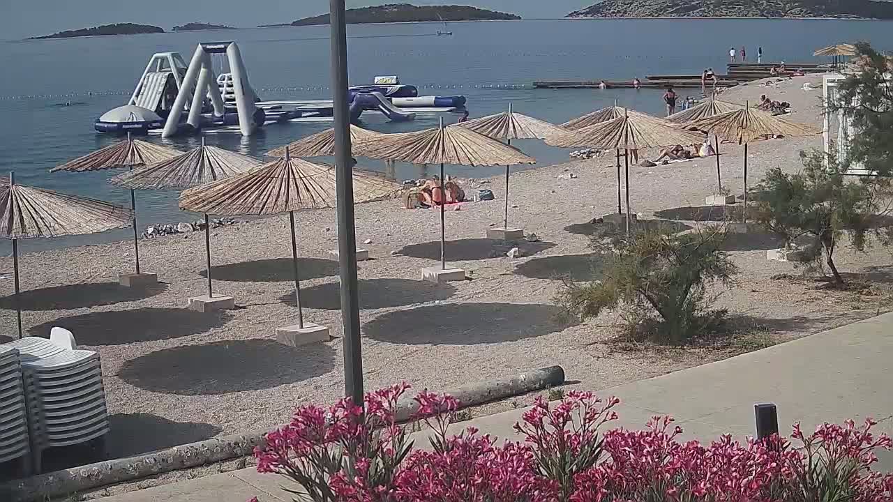 A sunny beach with numerous straw umbrellas, inflatable water toys in the water, and people relaxing on the sand.