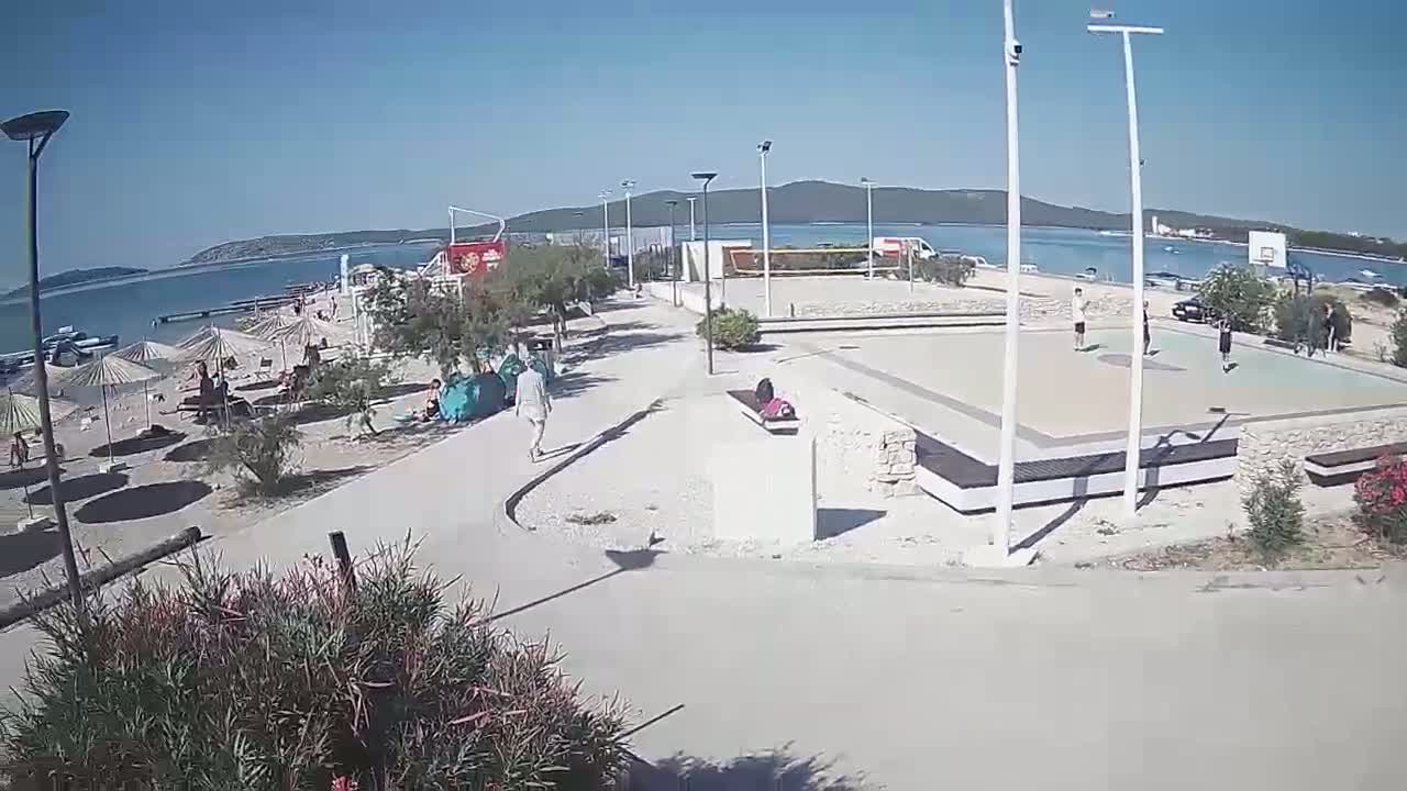 A beach scene on a sunny day shows people relaxing under umbrellas, others playing basketball on a nearby court, and a walkway leading from the beach to the court.