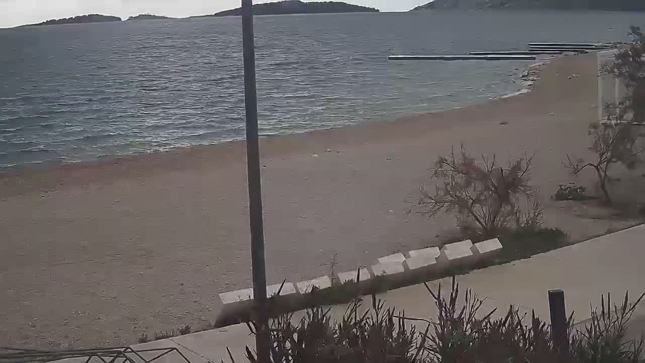 An overcast day overlooks a sandy beach with choppy water, a distant pier, and sparse, bare vegetation bordering a paved path with benches.