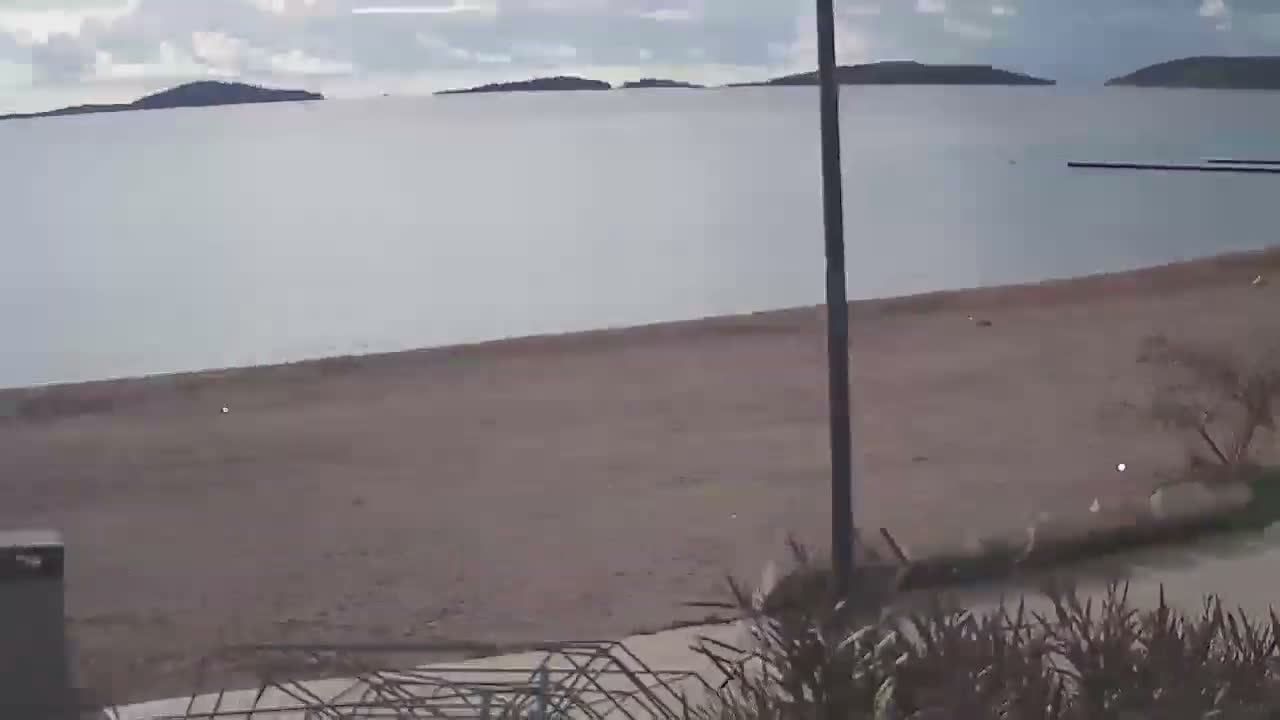 A wide, tranquil beach meets a calm body of water dotted with distant islands under an overcast sky, with a tall pole and some sparse vegetation visible in the foreground.