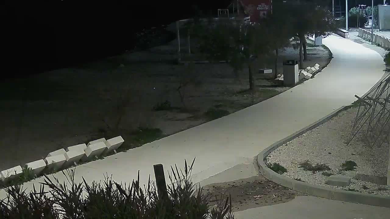 An outdoor scene at night reveals a brightly lit, winding paved pathway bordered by trees, dark ground, and white block structures on the left, and a gravel bed with a metal frame on the right, under clear skies.