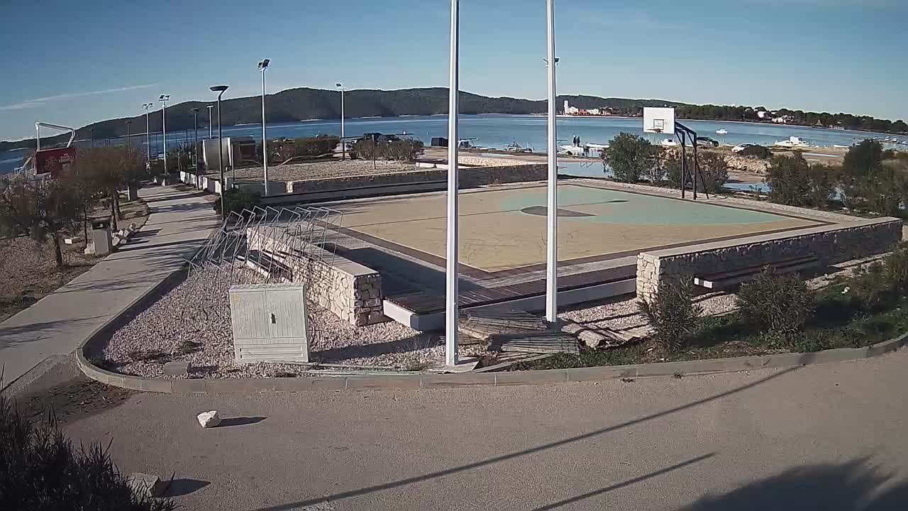 The image shows an empty outdoor basketball court and a paved walkway alongside a calm bay with distant green hills and a few buildings under a clear, sunny sky.
