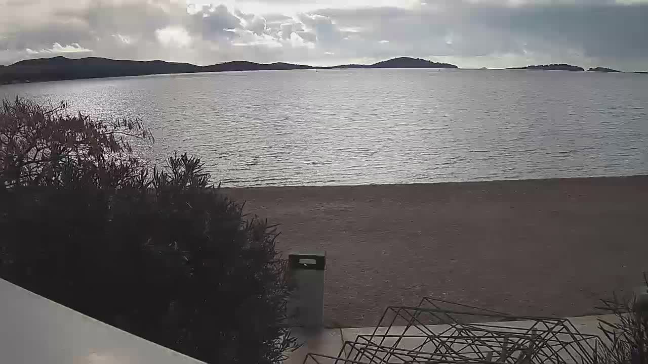 A tranquil bay with distant hills is viewed under a partly cloudy sky with muted sunlight, bordered by a narrow sandy beach and dark green foliage, a trash can, and geometric metal structures in the immediate foreground.
