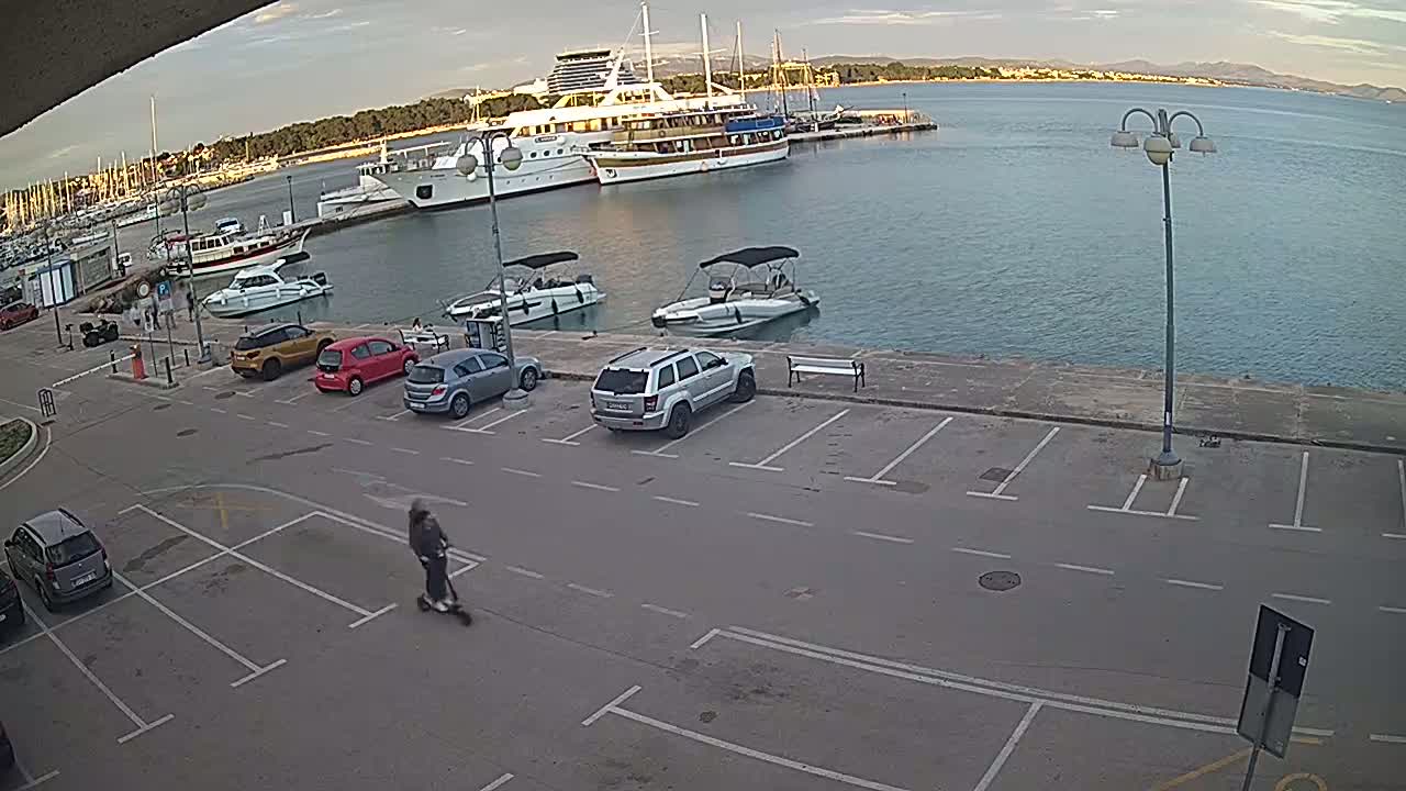 A person on a scooter rides past parked cars near a harbor filled with boats on a partly cloudy day.