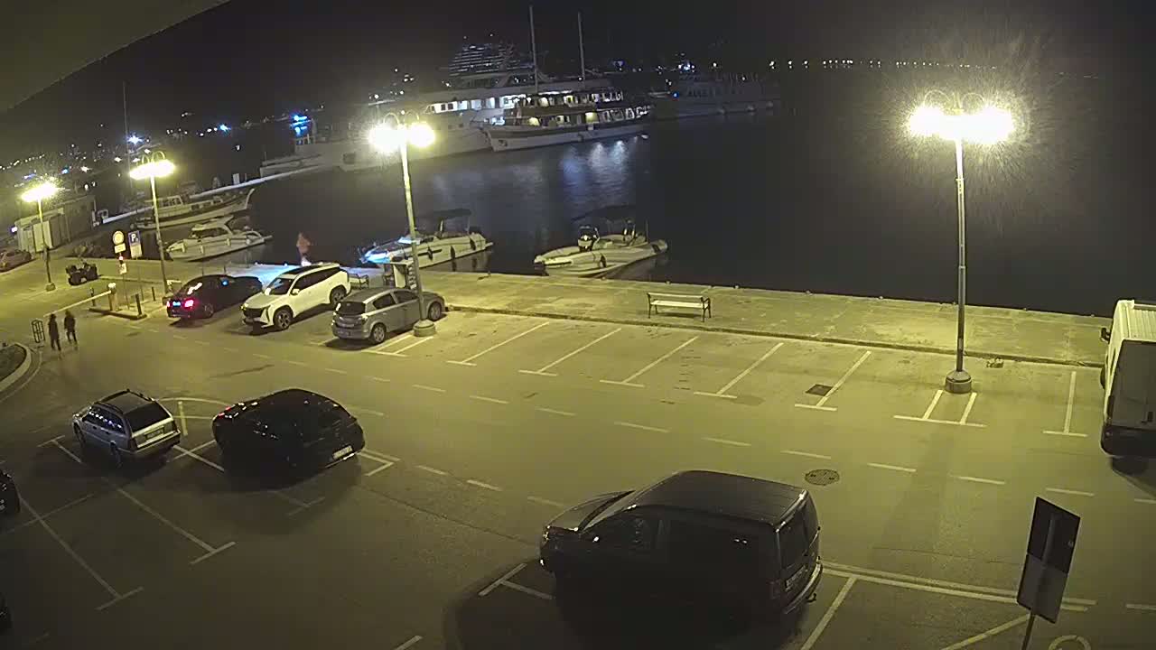 A nighttime view of a harbor with several cars parked in a lot next to a dock where boats and a large ship are moored.